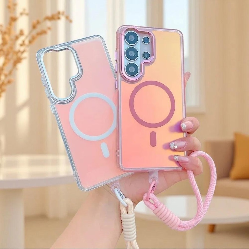 May include: Two rectangular smartphone cases with iridescent finishes. One case is clear with a white circular magnetic ring, and the other has a pinkish hue with a dark pink magnetic ring. Both feature attached woven wrist straps in light colours.