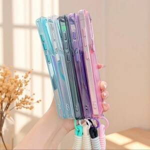 May include: A collection of five rectangular smartphone cases in translucent shades of light blue, clear, dark grey, purple, and pink. Each case features integrated side buttons and a small circular loop at the bottom attached to a matching coiled wrist strap.