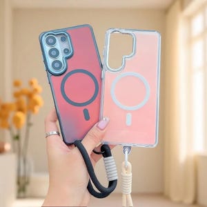May include: Two smartphone cases with magnetic charging rings on the back. One case is a dark red with a black camera frame and a black wrist strap. The other case is clear with a light pink tint, a white camera frame, and a white wrist strap.