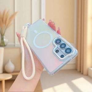 May include: A clear phone case with a soft iridescent gradient finish, featuring a white circular magnetic charging ring on the back and a matching white woven wrist strap attached to the side. The case has a raised silver camera lens frame.