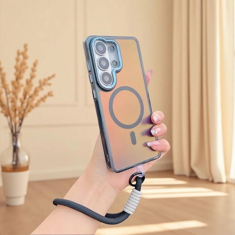 May include: A translucent, iridescent smartphone case with a blue camera frame and a circular magnetic charging ring on the back. The case features a dark blue wrist strap with a white ribbed connector attached to the bottom corner of the frame.