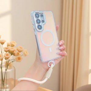 May include: A clear smartphone case with a circular magnetic charging ring on the back, featuring a multi-lens camera cutout and a thick, cream-coloured braided wrist strap attached to the bottom corner of the protective cover.