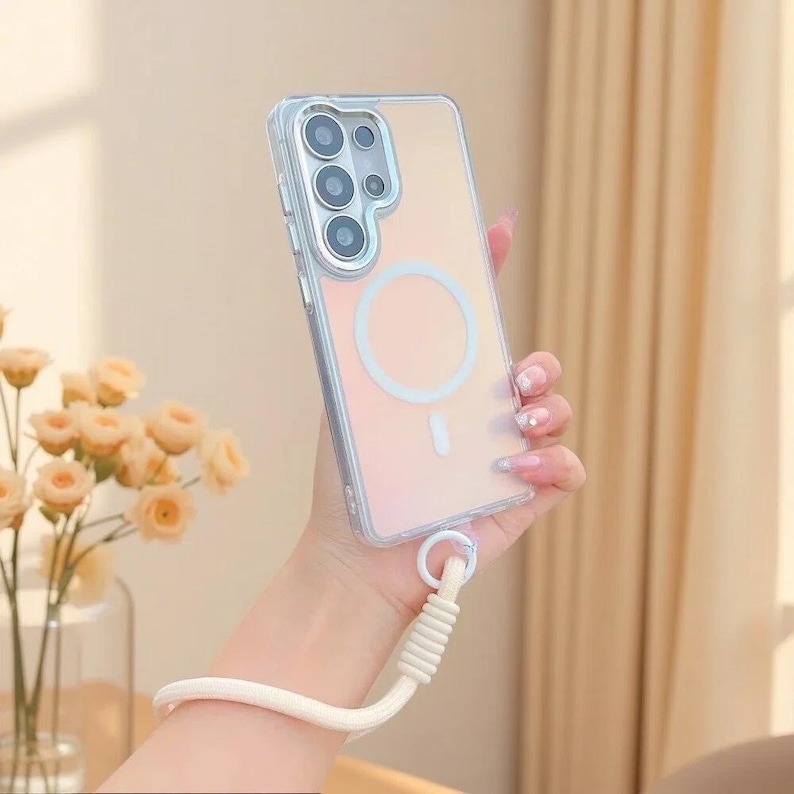 May include: A clear smartphone case with a circular magnetic charging ring on the back, featuring a multi-lens camera cutout and a thick, cream-coloured braided wrist strap attached to the bottom corner of the protective cover.