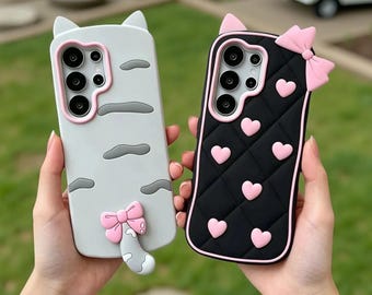 May include: Two smartphone cases with cat-ear tops. The left case is light grey with dark grey stripes and a pink bow on a tail. The right case is black with a quilted pattern, pink hearts, and a pink bow. Both feature pink camera cutouts.