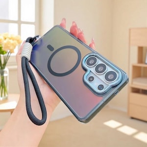 May include: A translucent, iridescent smartphone case with a gradient finish, featuring a circular magnetic charging ring on the back, a raised camera lens protector, and a black braided wrist strap with a white ribbed connector attached to the side.