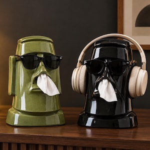 Puede incluir: Dos dispensadores de pañuelos de cerámica con forma de estatuas Moái. El de la izquierda es verde oliva y lleva gafas de sol negras. El de la derecha es negro brillante, lleva gafas de sol negras y auriculares beige.