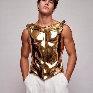 May include: A sleeveless, metallic gold chest piece with a highly reflective, liquid-like surface. The garment features a sculpted, form-fitting design that contours to the torso, with draped, folded edges at the shoulders and a pointed hem.
