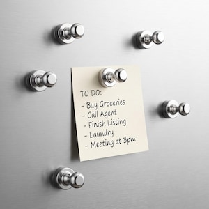 May include: Six silver-colored magnetic hooks attached to a metallic surface. One hook holds a square, off-white paper note with black text that reads: TO DO: - Buy Groceries - Call Agent - Finish Listing - Laundry - Meeting at 3pm.
