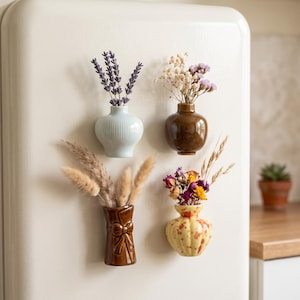 May include: Four small ceramic magnetic vases attached to a refrigerator surface. The vases include a light blue ribbed vase with lavender, a brown glossy vase with dried flowers, a brown bow-shaped vase with pampas grass, and a yellow speckled vase.