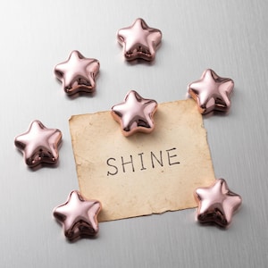 Könnte beinhalten: Sieben metallische roségoldene sternförmige Magnete, die um ein rechteckiges Stück gealtertes, cremefarbenes Papier angeordnet sind. Auf dem Papier steht das Wort SHINE in schwarzen Großbuchstaben. Die Magnete haben eine glatte, reflektierende Oberfläche.