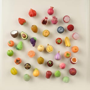Puede incluir: Una colección de imanes en miniatura con forma de fruta que presentan varios diseños realistas, incluyendo una fresa, aguacate, coco, uvas, granada, fruta del dragón, plátano, naranja, manzana y otras frutas tropicales y comunes.