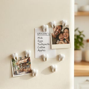 Puede incluir: Un conjunto de imanes blancos con forma de diente que sujetan una lista de la compra y dos fotografías en la superficie de un frigorífico. La lista incluye el texto Milk, Eggs, Toothpaste, Coffee y Apples. Los imanes tienen forma de molares.