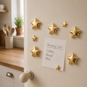 May include: A set of seven shiny gold star-shaped magnets arranged on a white surface. A rectangular white note is attached to the surface, featuring the handwritten text: Grocery List: Coffee, Bread, Milk. The magnets vary in size and shape.