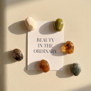 May include: A rectangular white card with the black printed text BEAUTY IN THE ORDINARY, surrounded by six smooth, polished, irregularly shaped stones in various colors including cream, olive green, amber, dark brown, and greyish-blue.