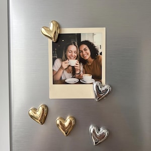Könnte beinhalten: Ein quadratisches Foto von zwei lächelnden Personen mit weißen Kaffeetassen, das mit fünf dreidimensionalen herzförmigen Magneten an einer gebürsteten Metalloberfläche befestigt ist. Drei Magnete sind goldfarben und zwei silberfarben, alle glatt.