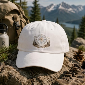 May include: A white baseball cap featuring a brown embroidered graphic of a compass centered above a landscape of pine trees and mountain peaks. The cap has a structured crown, a curved brim, and eyelets for ventilation on the top panels.