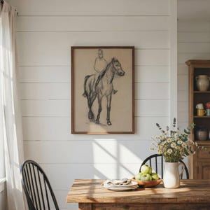 May include: A framed charcoal sketch on beige paper depicting a person riding a horse, mounted on a white shiplap wall. The artwork features loose, expressive lines capturing the form of the rider and the horse in a standing position.