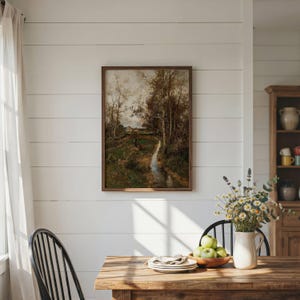 May include: A framed landscape painting depicting a stream flowing through a wooded area with trees and a small building in the distance. The artwork is displayed on a white horizontal plank wall above a wooden table with a vase of flowers.