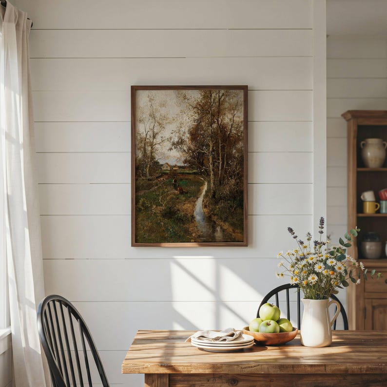May include: A framed landscape painting depicting a stream flowing through a wooded area with trees and a small building in the distance. The artwork is displayed on a white horizontal plank wall above a wooden table with a vase of flowers.