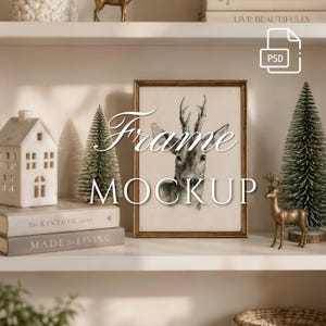 May include: A vertical rectangular picture frame with a thin gold border containing a black and white sketch of a deer head. The frame is displayed on a shelf alongside a white ceramic house, two small bottle brush trees, and a gold deer figurine.