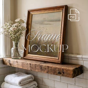 May include: A rectangular wooden picture frame with a gold inner trim containing a landscape painting, resting on a rustic wooden shelf. Next to the frame is a clear glass vase filled with white flowers. The text "Frame MOCKUP" is visible.