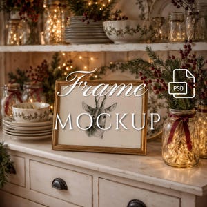 May include: A rectangular gold-toned picture frame containing a black and white sketch of a deer head. The frame sits on a white wooden surface surrounded by glass jars filled with fairy lights, stacks of white bowls, and sprigs of red berries.