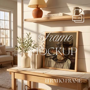 May include: A square wooden picture frame mockup featuring a still life painting of a glass, a plate with food, and a white cloth. The frame is displayed on a wooden console table next to a cream-colored ceramic vase with green leaves.