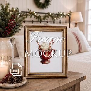 May include: A gold-toned picture frame containing an illustration of a red mug filled with whipped cream and candy canes. The text "Frame MOCKUP" is overlaid on the image. The frame sits on a wooden surface next to a white pitcher and pinecones.