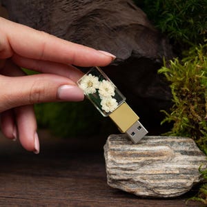 Può includere: Una chiavetta USB rettangolare con un involucro in resina trasparente che racchiude tre piccoli fiori bianchi essiccati. Il dispositivo presenta un connettore in metallo color oro e una presa USB argentata standard alla base della resina.