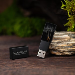 Könnte beinhalten: Ein schwarzer USB-Stick mit einer Pusteblumen-Grafik auf dem Gehäuse und einer separaten Kappe mit dem weißen Schriftzug MOONSTONE PHOTOKRAFTFILM. Der Stick hat einen Metallanschluss und ein rechteckiges schwarzes Gehäuse mit glatter Oberfläche.