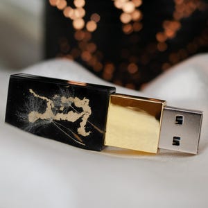Puede incluir: Una unidad flash USB rectangular negra con un diseño de cabeza de semilla de diente de león dorado en la carcasa, conectada a una base cuadrada de tono dorado pulido y un conector USB de metal plateado estándar.