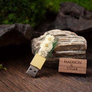 May include: A USB flash drive featuring a clear resin cap embedded with small white dried flowers and a gold-toned metal base. Beside it lies a rectangular wooden cap engraved with the text MADISON & TYLER in a dark brown serif font.