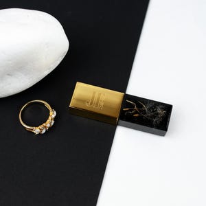 Può includere: Un anello dorato con diverse piccole pietre bianche è posizionato accanto a una pietra bianca liscia e a una scatola rettangolare. La scatola ha un coperchio dorato inciso con le lettere JE e una base nera contenente un tarassaco.