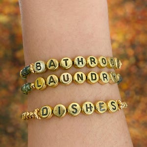 May include: Three gold beaded bracelets spelling out "BATHROOM", "LAUNDRY", and "DISHES" in black letters. The bracelets are worn on a wrist against a blurred, autumnal background.