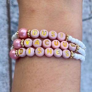 May include: Stacked bracelets with pink and white beads. The top bracelet has pink pearl beads and gold accents. The middle bracelet has pink letter beads that spell "DUST VACUUM DISHES". The bottom bracelet is white.
