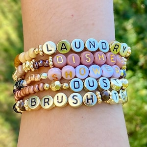 May include: A stack of beaded bracelets with gold and colorful beads. Each bracelet features letter beads spelling out words like "LAUNDRY", "DISHES", "SHOWER", "DUST", and "BRUSH". The bracelets are stacked on a wrist.