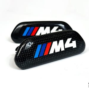 May include: Two black carbon fibre automotive emblems featuring the M4 logo in white text next to three diagonal stripes in light blue, dark blue, and red. The emblems have a glossy finish and a curved, rectangular shape with rounded corners.