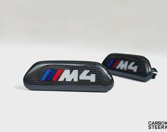 May include: Two automotive interior trim pieces featuring a black carbon fiber texture. Each piece displays the M4 logo in white lettering, accompanied by three slanted stripes in blue, purple, and red. The text CARBON STEERA is visible.