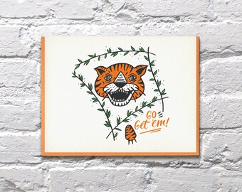 May include: A greeting card with an orange and black tiger face illustration surrounded by green leaves. The text "GO get 'em!" is written in orange below the tiger.