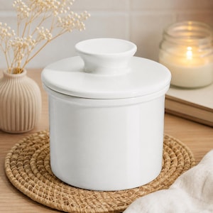 May include: A white ceramic butter crock with a smooth, glossy finish and a matching lid featuring a central knob handle. The cylindrical container has a clean, simple design with straight sides and a flat base, providing a functional kitchen storage piece.