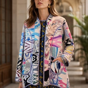 May include: A long-sleeved, quilted jacket featuring a multi-coloured patchwork pattern with shades of blue, pink, yellow, and black. The jacket has a round neckline, a front button closure, and two large patch pockets, all finished with light purple trim.