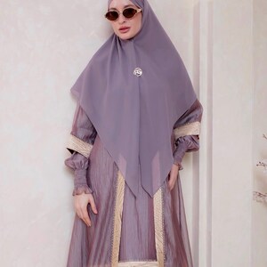 May include: A mauve hijab paired with a matching long-sleeved dress featuring a textured, crinkled fabric. The dress includes beige panels with a subtle pattern along the front and sleeves, finished with smocked cuffs and a circular gold-tone brooch.