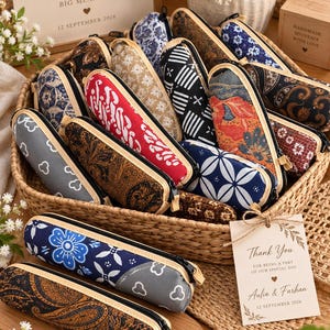 Op de afbeelding: Een verzameling cilindervormige etuis van stof met diverse batik- en geometrische patronen in kleuren zoals rood, blauw, zwart, bruin en grijs, elk voorzien van een zwarte ritssluiting en een lichte rand, gepresenteerd in een gevlochten mand.
