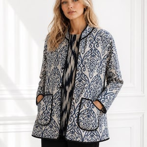 May include: A quilted jacket featuring a beige base with a dark blue damask pattern, a stand-up collar, long sleeves, and two front patch pockets. The jacket is finished with black piping along the edges and front closure with visible buttons.