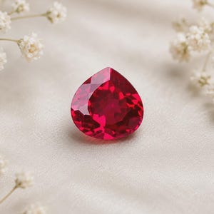 May include: A pear-shaped, faceted gemstone with a deep, vibrant red color. The stone features a pointed top and a rounded base, with numerous geometric facets that reflect light across its surface, creating a brilliant, sparkling appearance.