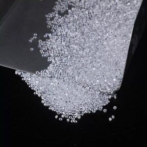 Può includere: Una grande collezione di piccole pietre preziose trasparenti e sfaccettate sparse su una superficie scura. Le pietre vengono versate da un misurino trasparente, creando un denso mucchio di cristalli scintillanti, incolori e rotondi.