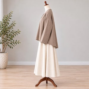 May include: A taupe long-sleeved top with a V-neckline paired with a cream-colored A-line midi skirt. The ensemble is displayed on a mannequin, showcasing a loose-fitting silhouette with a high-low hemline on the top and a flared skirt.