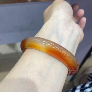 Puede incluir: Una pulsera tipo brazalete lisa y translúcida que presenta un degradado de tonos naranja cálido y ámbar intenso. El accesorio circular tiene un acabado pulido y brillante que capta la luz, mostrando un diseño de banda sólida y continua con bordes redondeados.