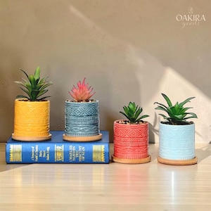 May include: Four cylindrical ceramic planters with embossed geometric patterns, each resting on a circular bamboo saucer. The planters are yellow, blue, red, and light blue, containing small artificial succulents. The text OAKIRA goods is visible.