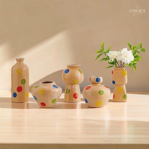 Pode incluir: Cinco vasos de cerâmica bege de várias formas e tamanhos, cada um decorado com bolinhas coloridas pintadas à mão em vermelho, azul, verde e amarelo. Um vaso contém um buquê de flores brancas. O texto OAKIRA goods é visível no canto.